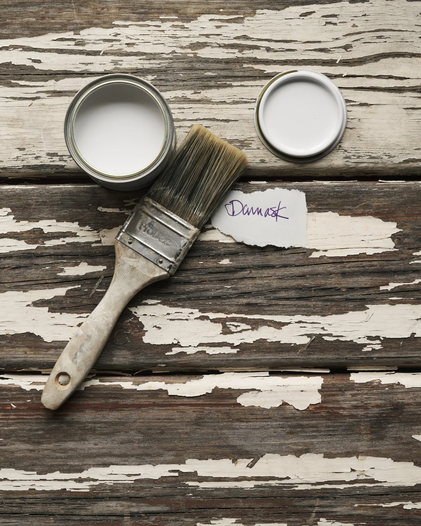 An overhead view of an open paint pot and a worn brush on a rustic tabletop. The off-white colour inside is labelled 'Damask'.