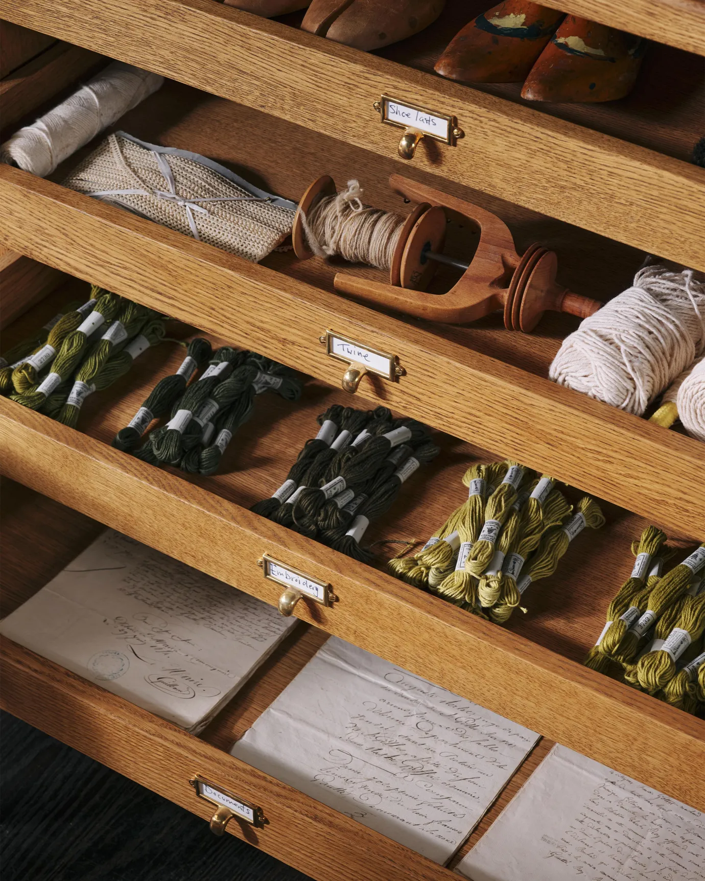 An oblique view partially open oak drawers, fitted with small cardframe pull handles in a warm burnished brass finish and a handwritten paper labels that read 'Twine', 'Embroidery', and 'Documents'.