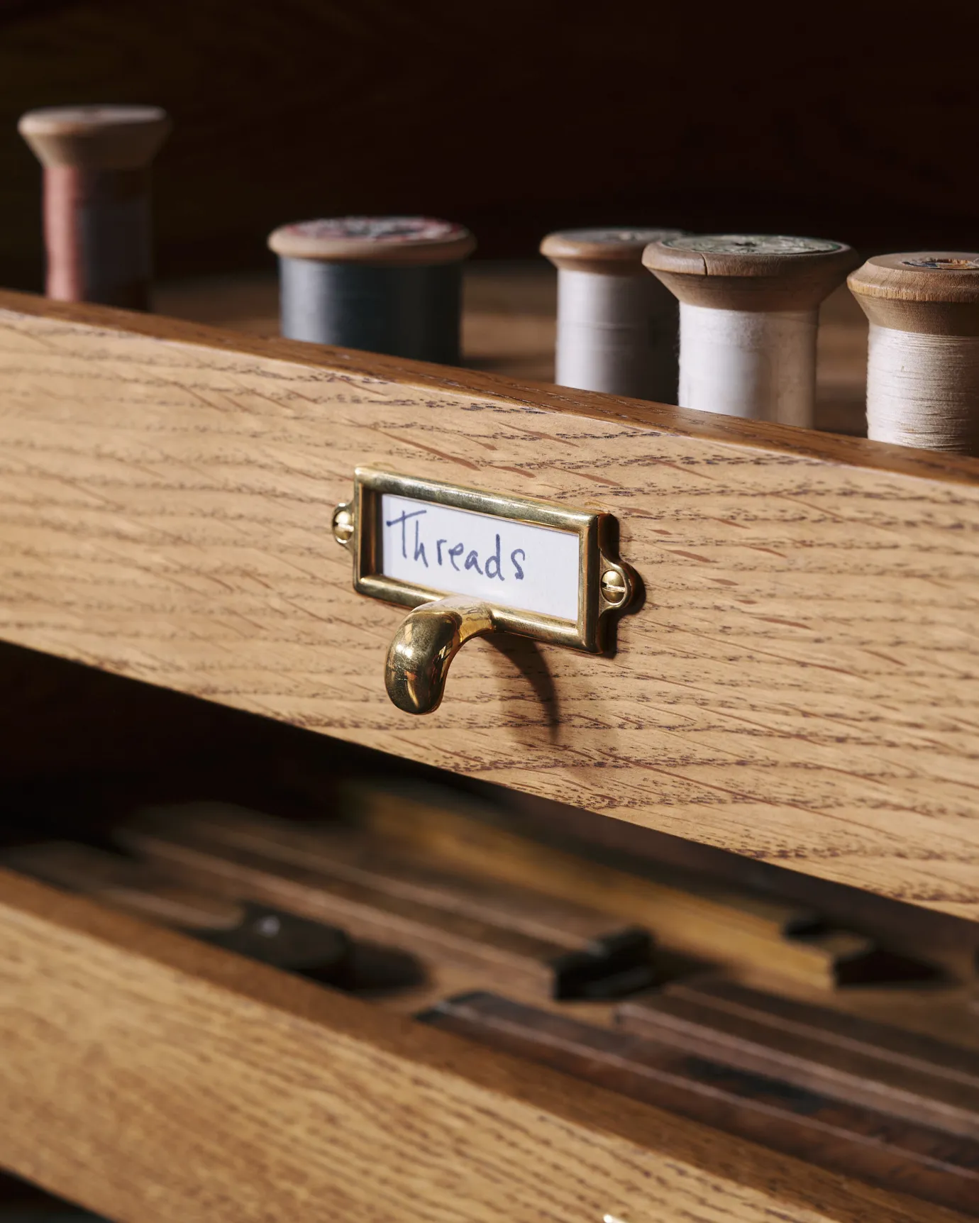 A close-up oblique view of a small cardframe pull handle in a warm burnished brass finish and a handwritten paper label that reads 'Threads', fitted to an oak drawer front partially concealing spools of coloured thread.