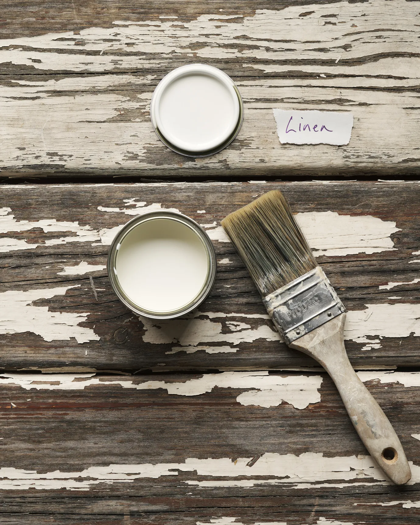 An overhead view of an open paint pot and a worn brush on a rustic tabletop. The light cream colour inside is labelled 'Linen'.