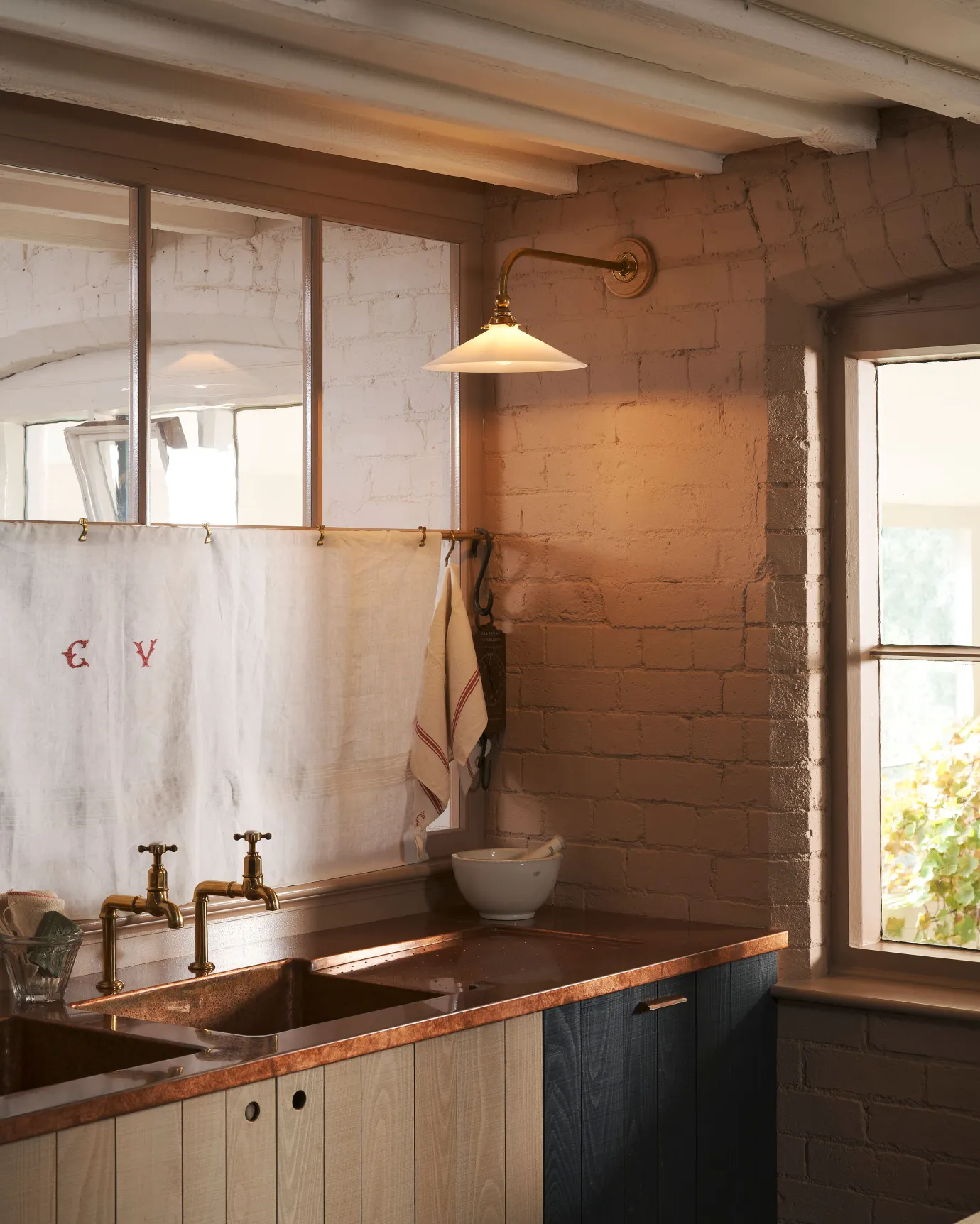 An oblique view of a large Victorian-inspired wall light with a brass arm and a milk glass shade fixed to a pink brick wall over modern rustic kitchen with an aged copper worktop.