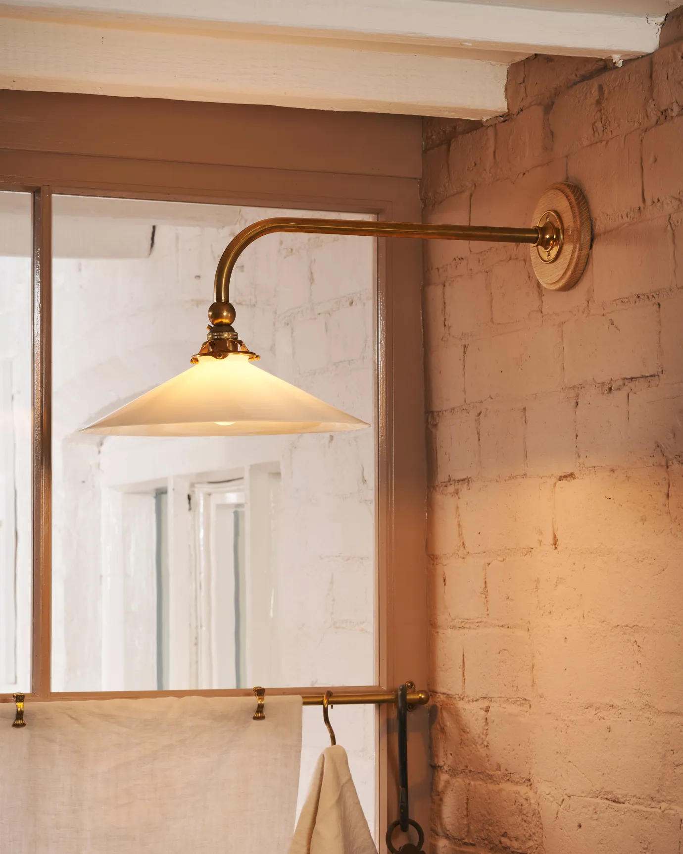 A close-up oblique view of a large Victorian-inspired wall light with a brass arm and a milk glass shade fixed to a pink brick wall next to a glazed partition.
