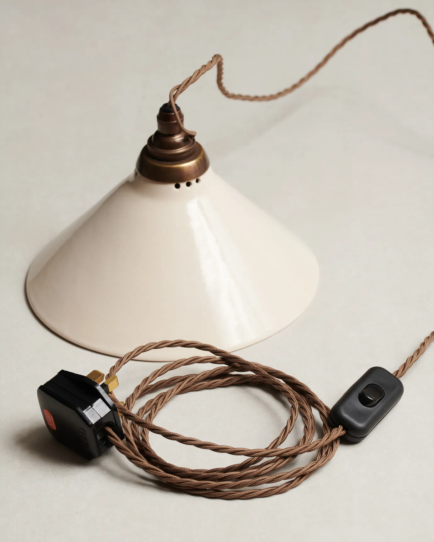 A small plug-in light with a cream glazed shade, brown twisted fabric flex cord, brass fittings and a black plastic switch and plug.