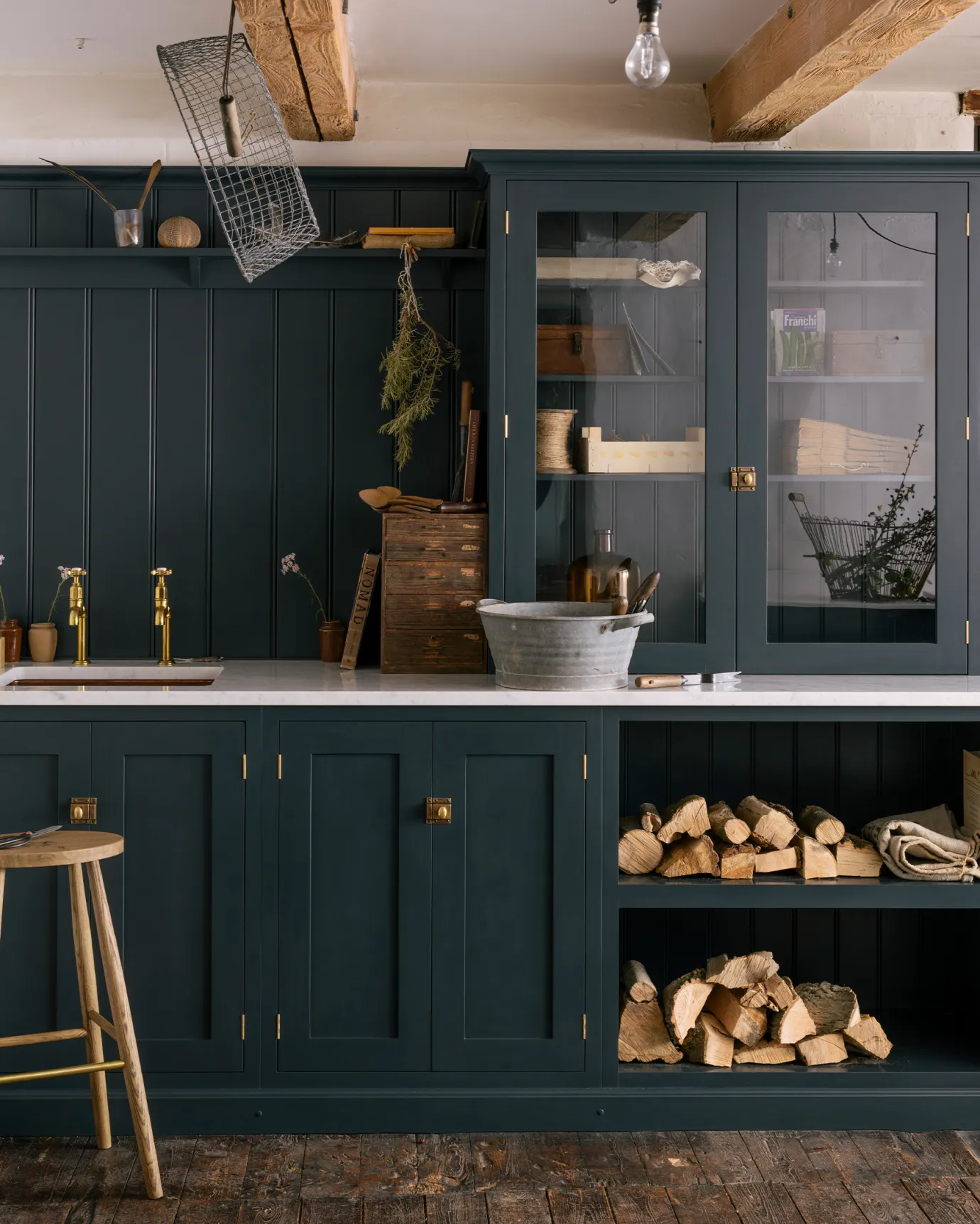 A straight-on view of a simple Shaker-style utility kitchen with dark blue painted cupboards, aged brass taps, tongue and groove-style panelling and a glazed countertop cupboard. The styling is rustic with logs piled in an open cupboard and a galvanised steel tub on the worktop.