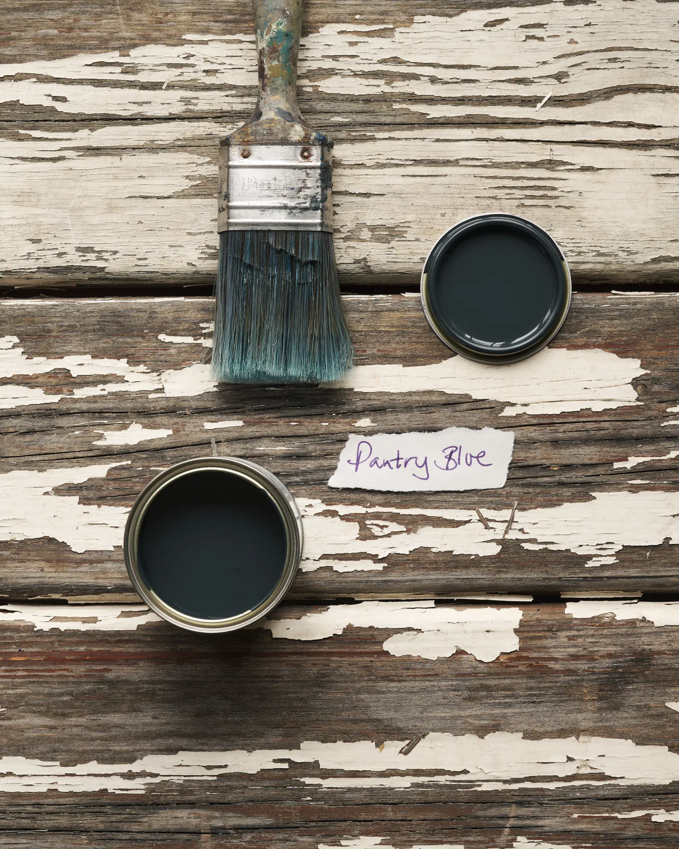An overhead view of an open paint pot and a worn brush on a rustic tabletop. The dark blue colour inside is labelled 'Pantry Blue'.