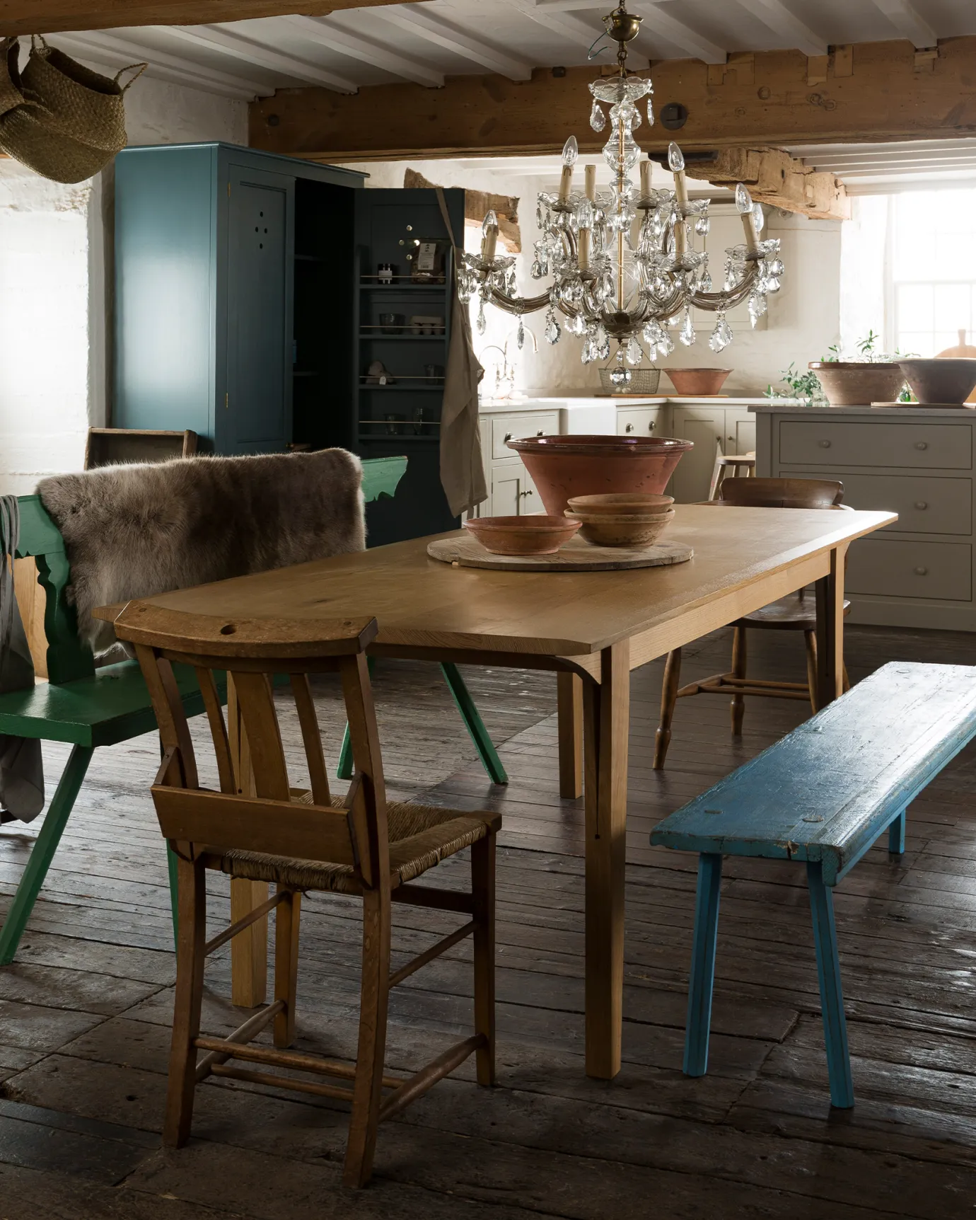 An oblique view of a mid-century modern-style oak dining table in a rustic cottage with dark worn floorboards and white brick walls.