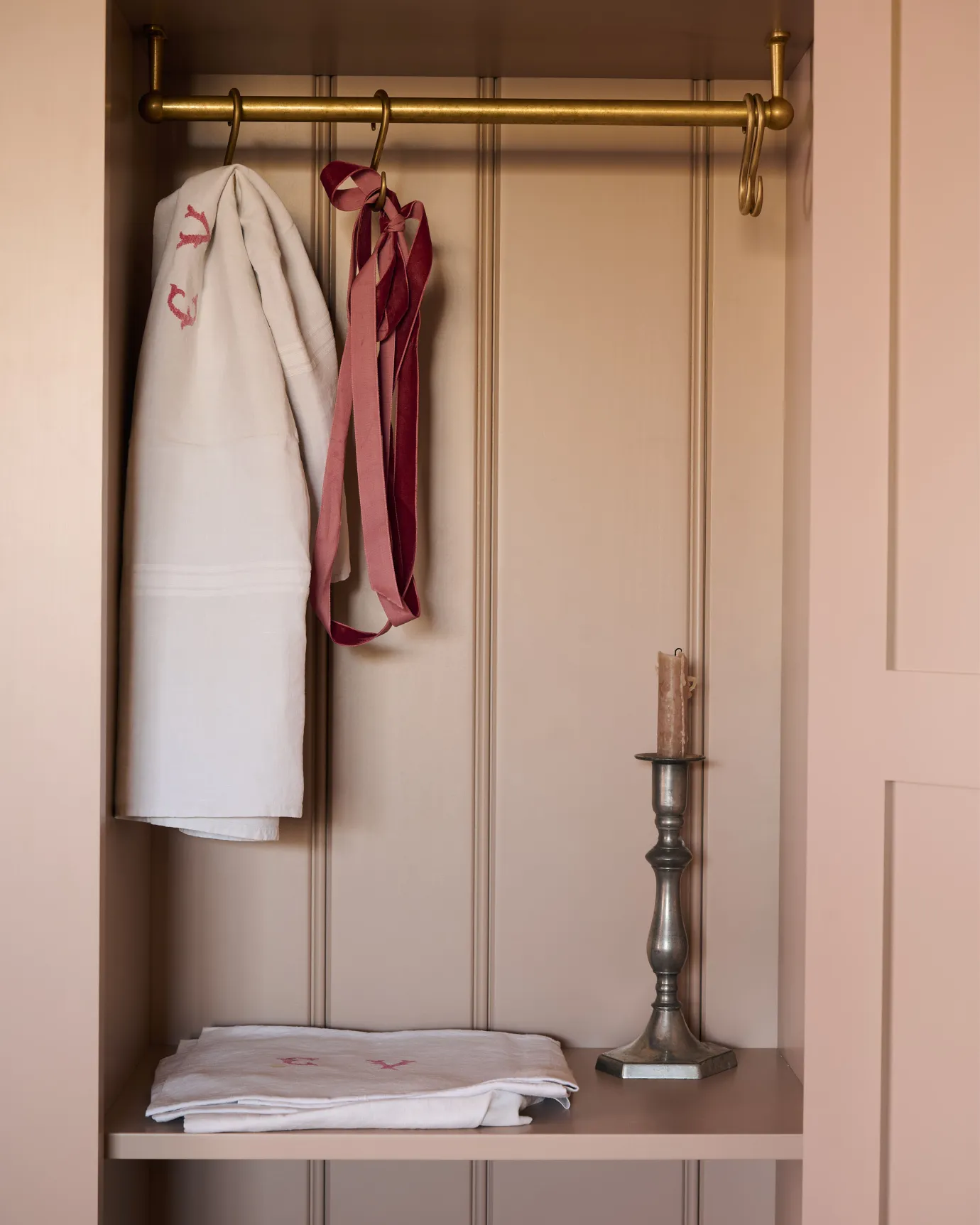 Looking into a Victorian-style pantry cupboard, painted inside and out in a soft pink colour. Inside the cupboard, there is a brass hanging rail with vintage linens and a bronze candlestick.