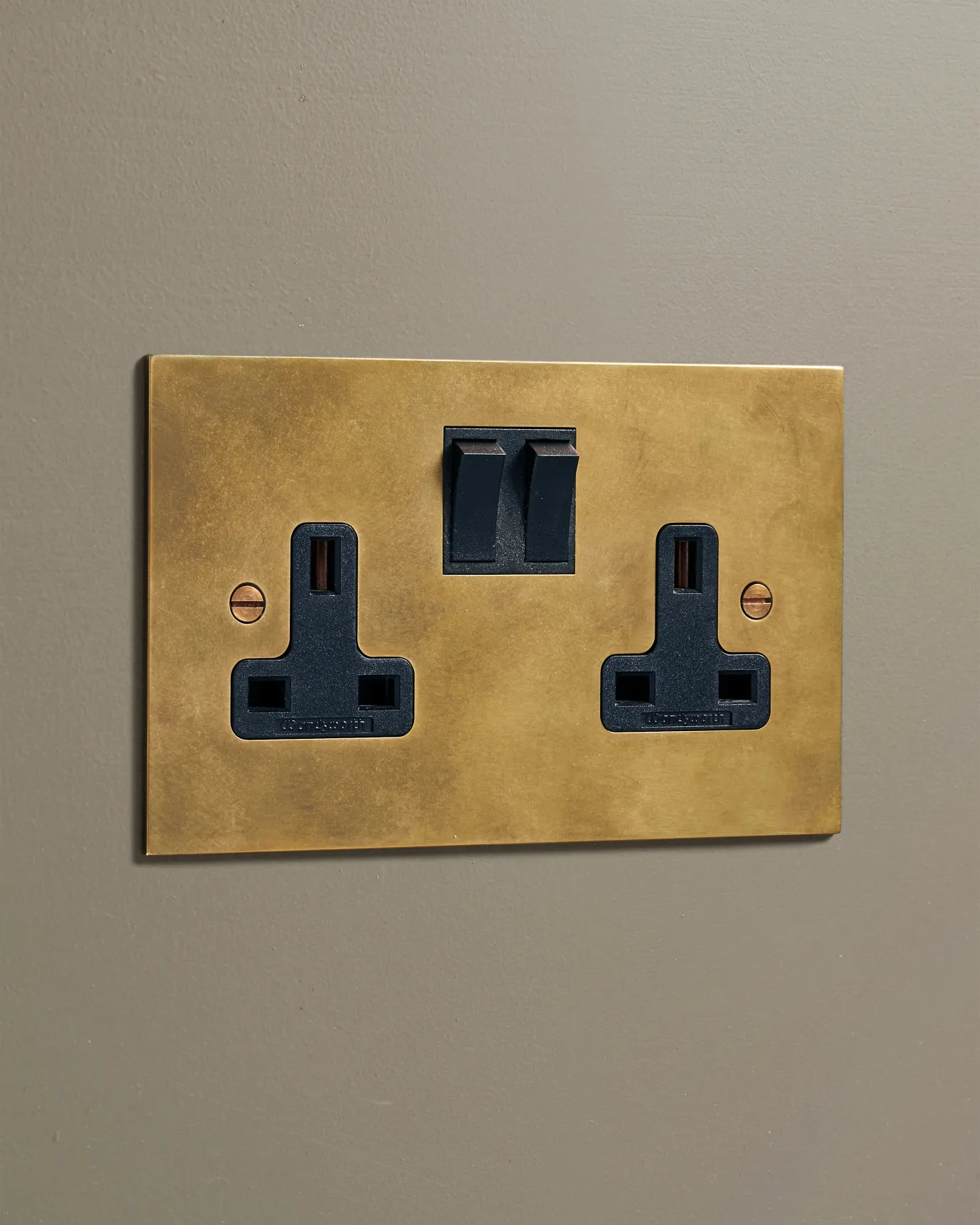 An oblique view of a classic 13 amp twin plug socket in a warm burnished brass finish with black plastic switches.