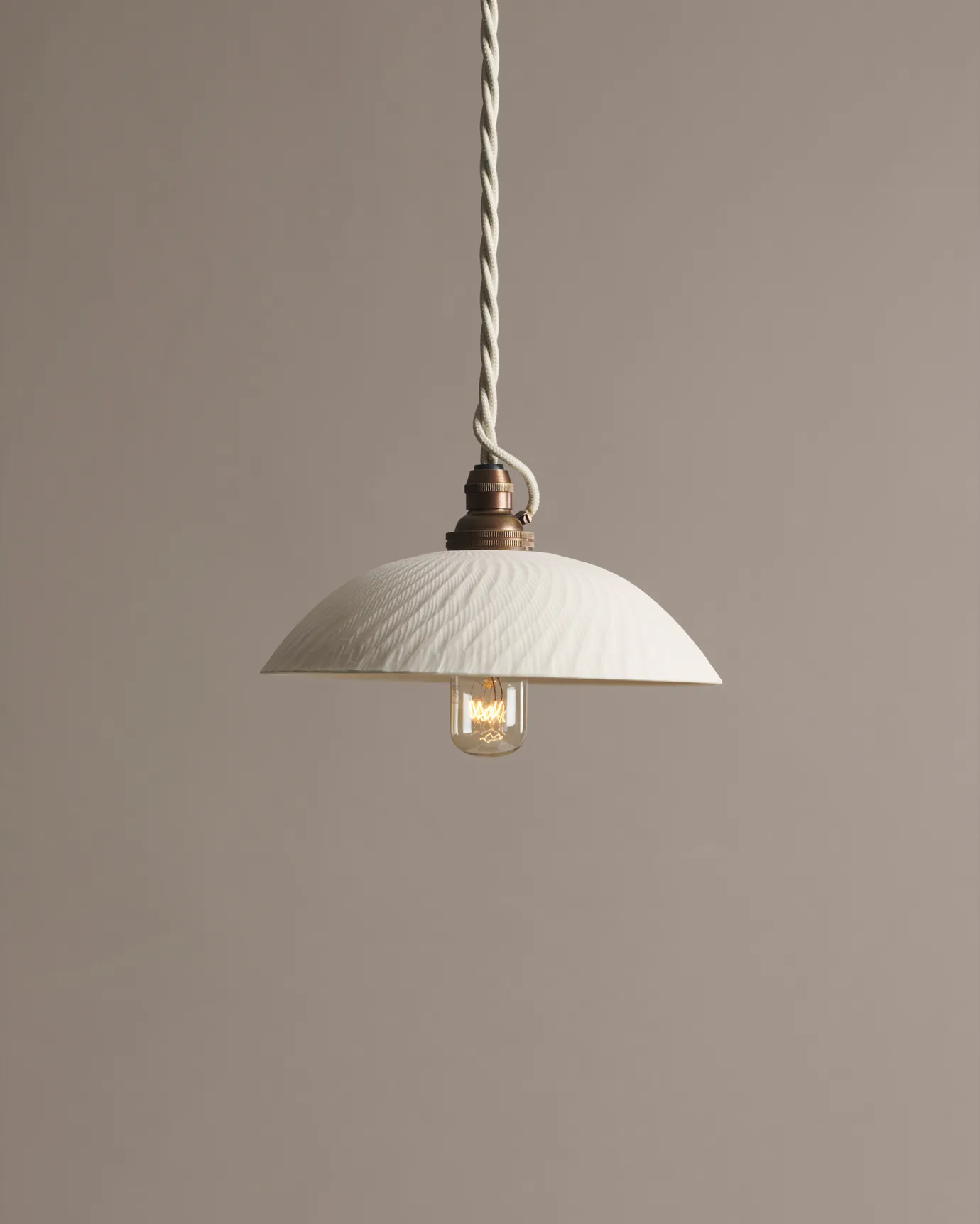A small chattered skullcap porcelain pendant light with an unglazed finish, an aged brass E27 lampholder and a white twisted fabric flex cord.
