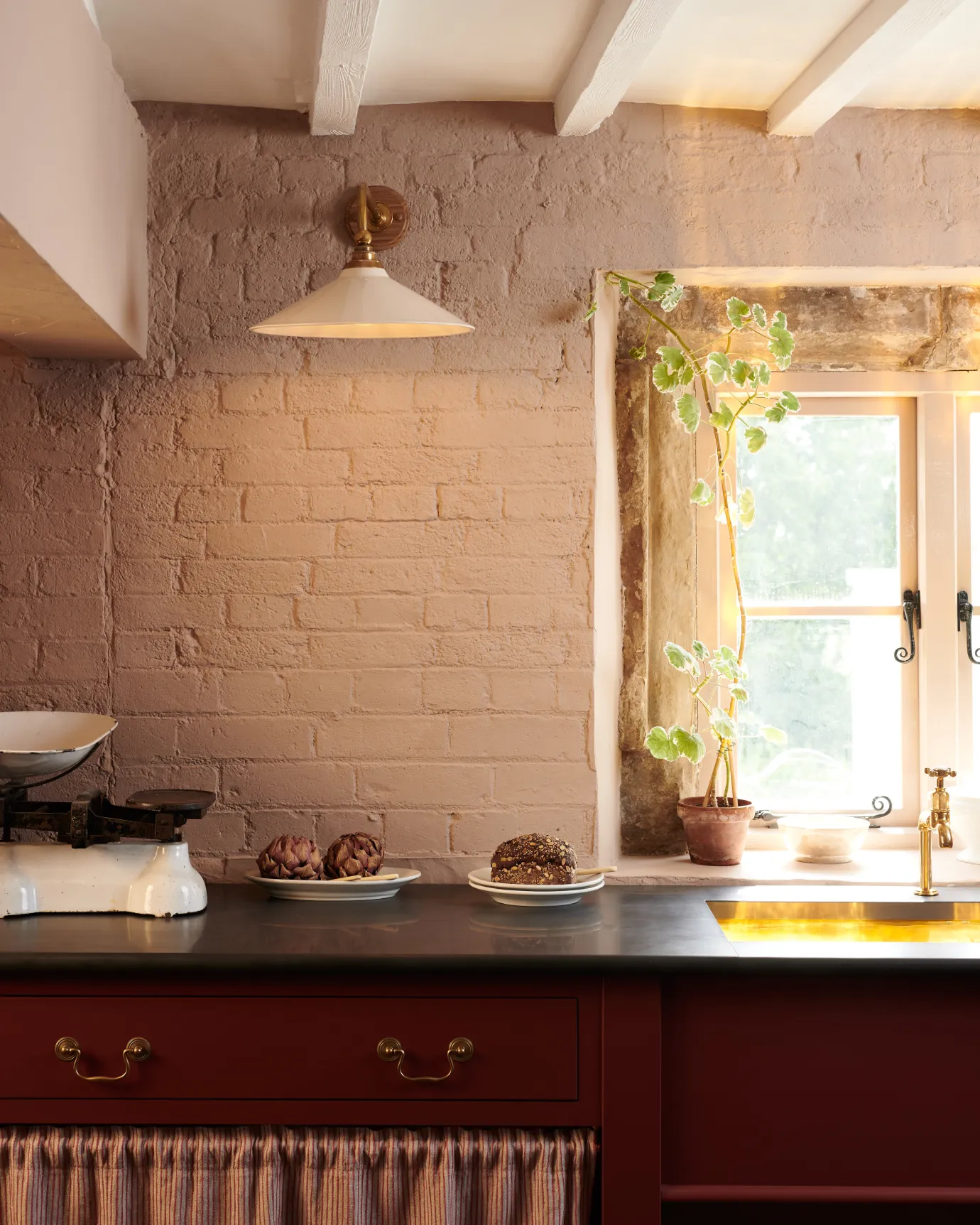 A straight-on view of a large Victorian-inspired wall light with a brass arm and a cream glazed ceramic shade fixed to a pink painted brick wall above a slate worktop and red painted cabinetry.