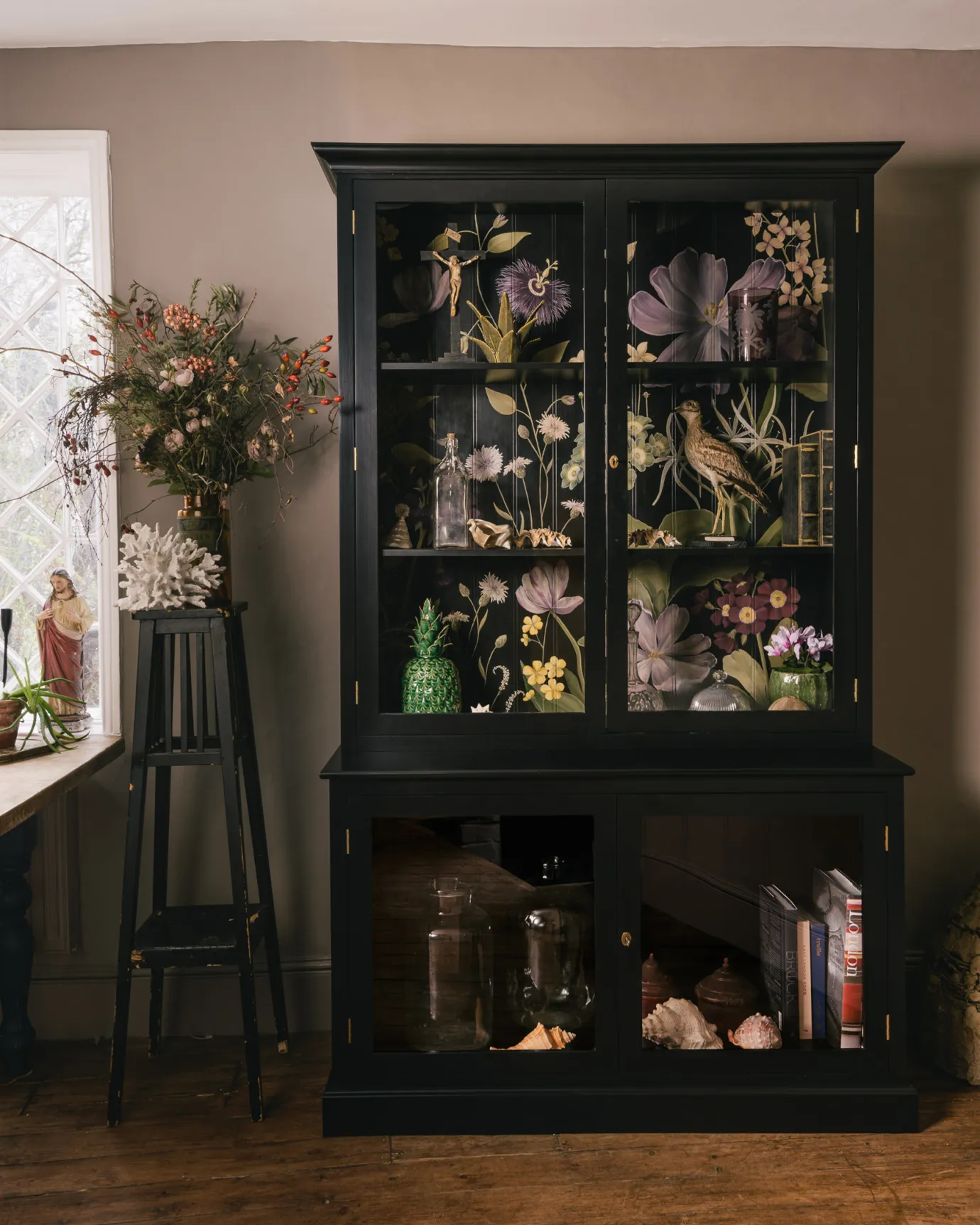 A tall museum-style display cabinet with glazed doors, painted black inside and out and with an elegant and bustling painted floral design on the tongue and groove back panel.