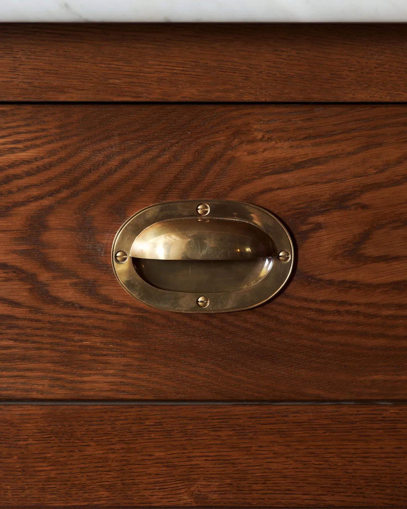 A straight-on view of a Victorian-inspired oval-shaped cup handle with an authentic and warm burnished brass finish and a recessed backplate, fitted to a dark stained oak drawer front.