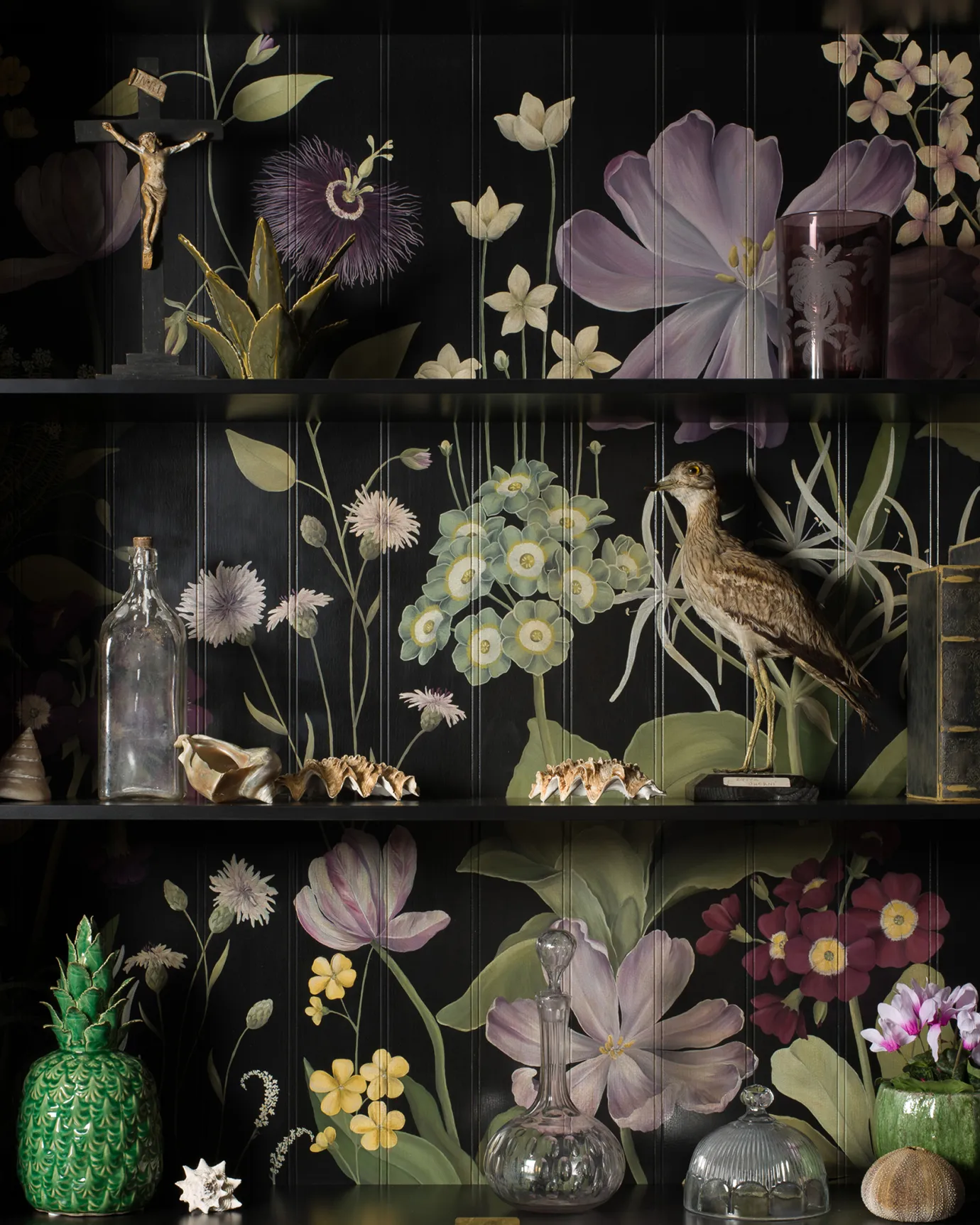 The interior of a black display cabinet with an elegant and bustling painted floral design on a tongue and groove back panel. The shelves are full of vintage oddities and taxidermy.