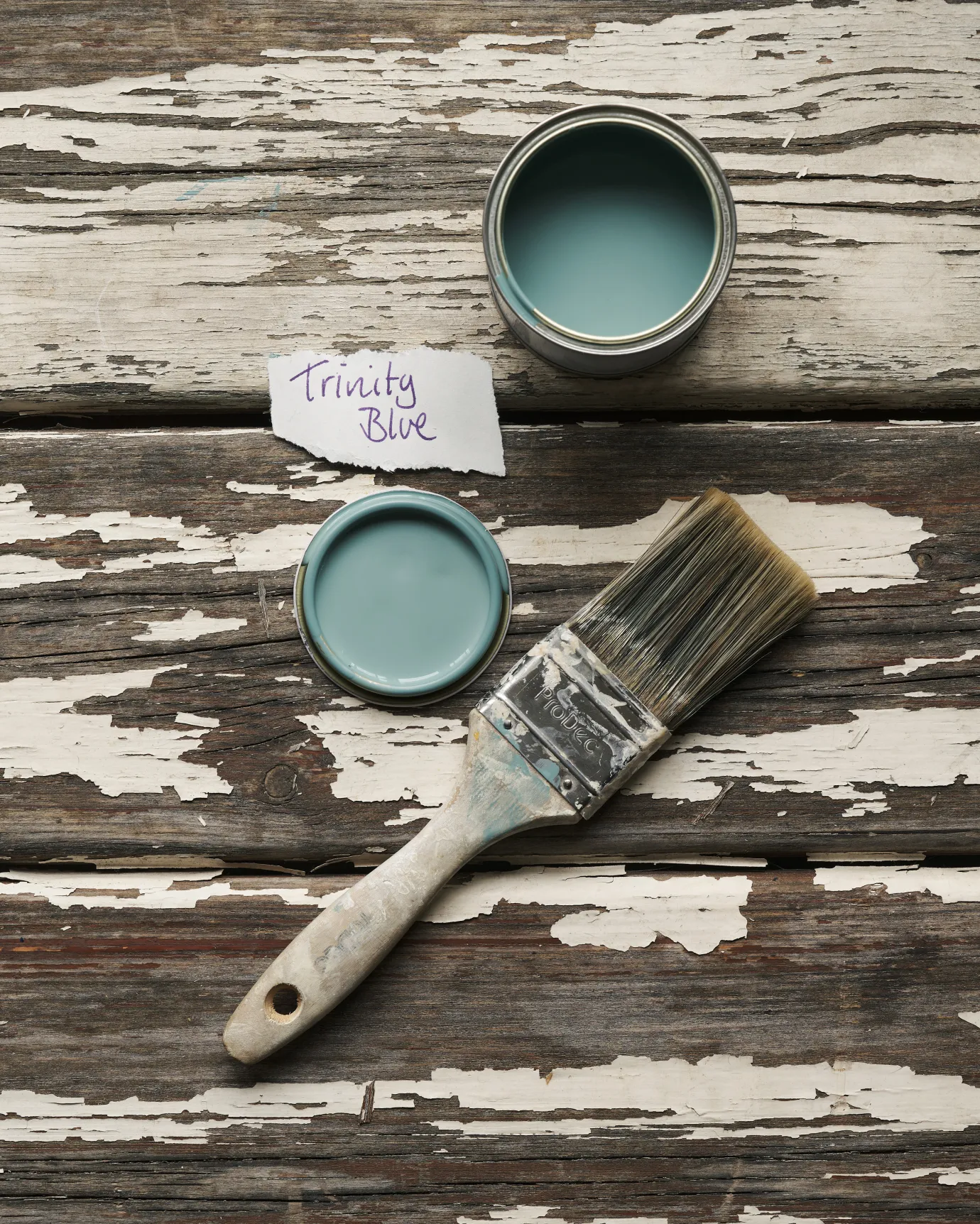 An overhead view of an open paint pot and a worn brush on a rustic tabletop. The duck-egg blue colour inside is labelled 'Trinity Blue'.